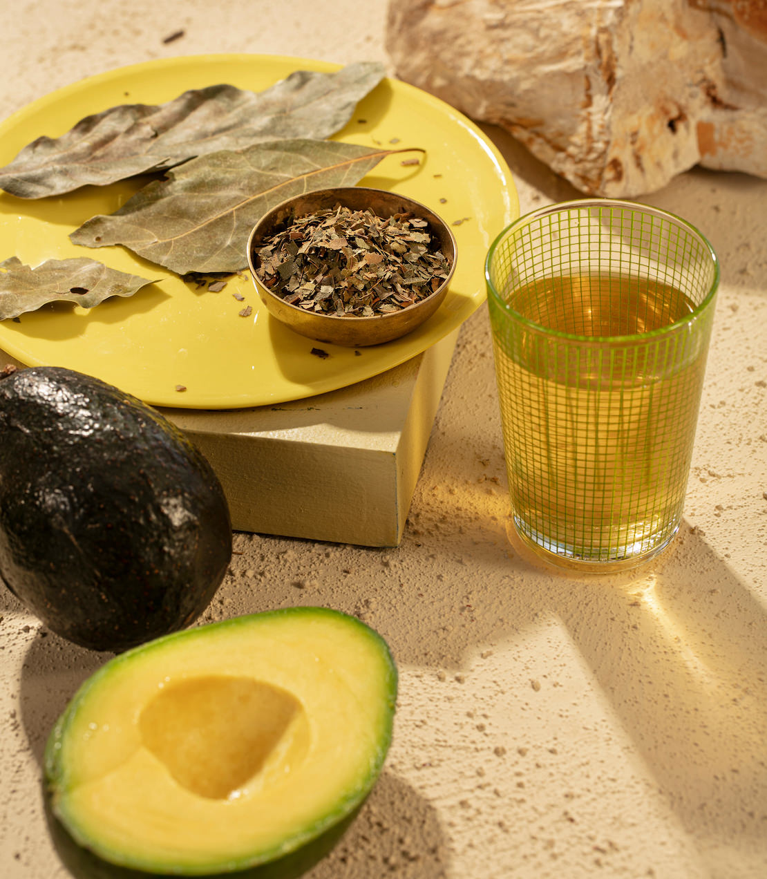 Avocado leaves and a sliced avocado beside a glass of herbal tea on a yellow plate, showcasing natural wellness.