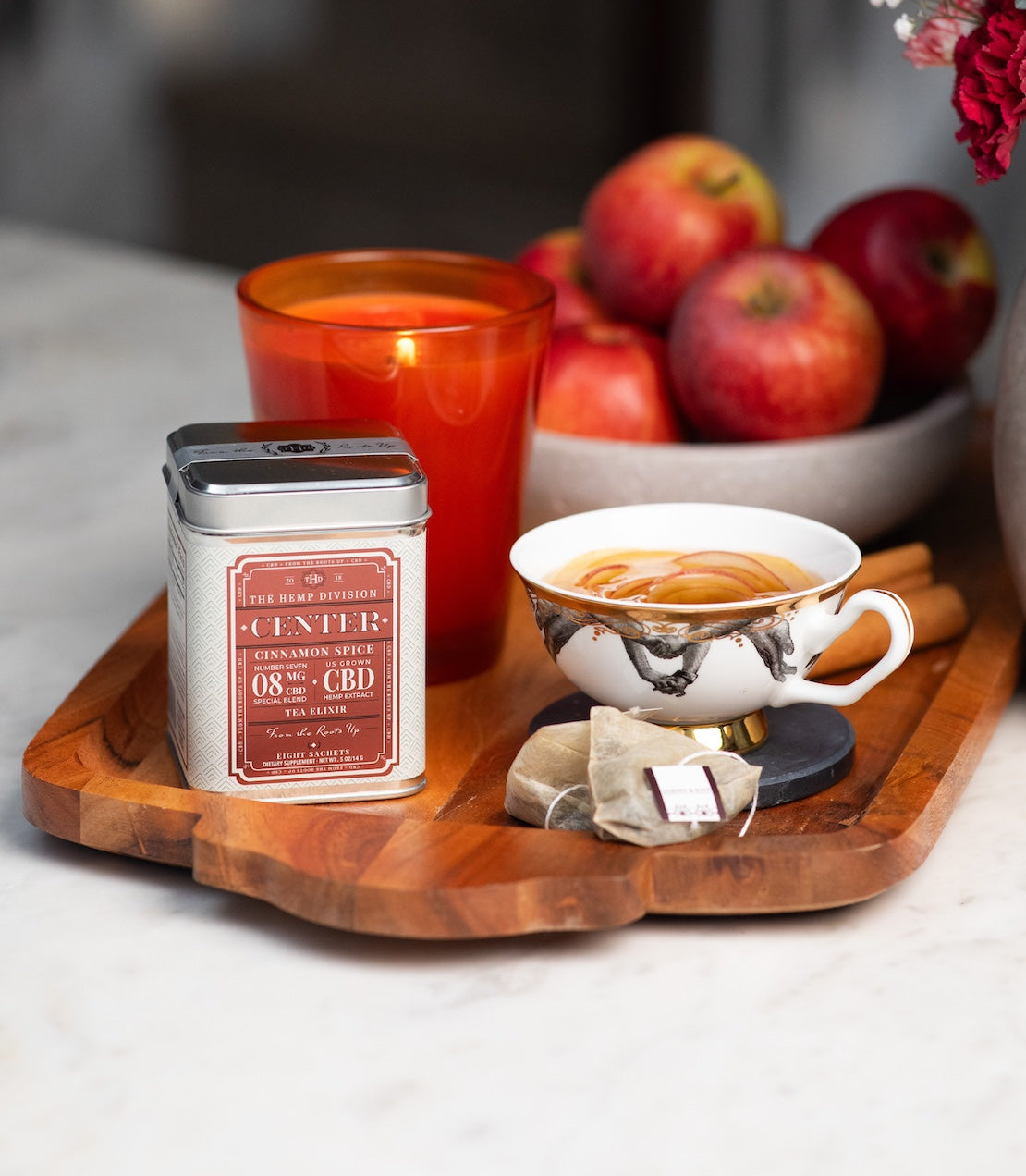 Center Cinnamon Spice 8 MG CBD tea on a marble table on top of a wooden tray. Teacup and tea sachets are next to the tin. In the background there's fall decor with a red candle and apples.