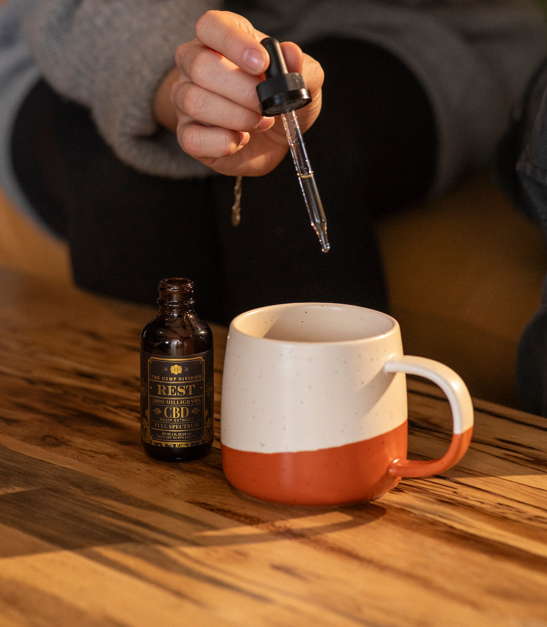 Extra Strength CBD Hemp Extract Tincture being added to a warm beverage in a glass mug, with cinnamon and lemon in the background.