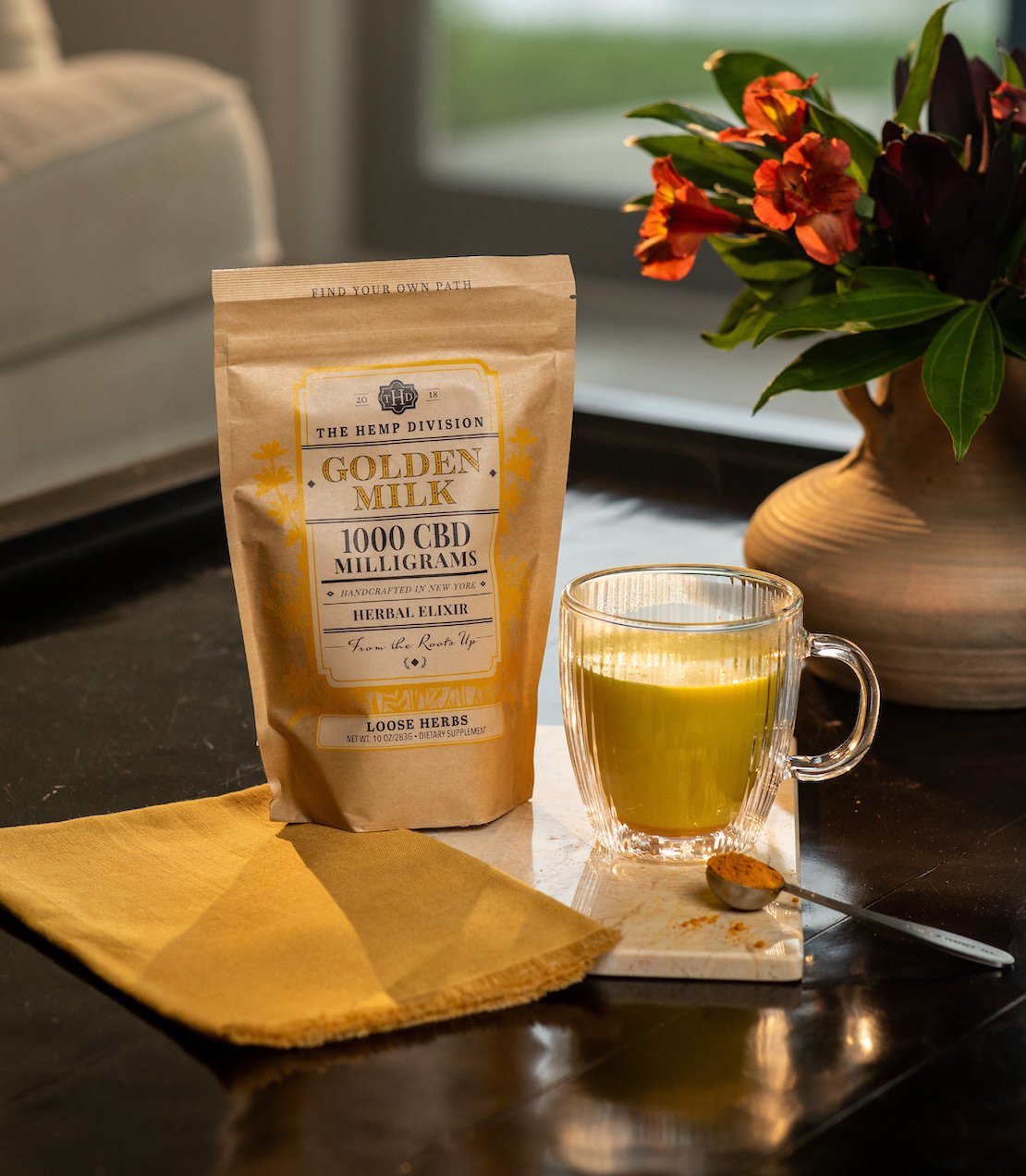 Golden Milk 1000 MG CBD Elixir on a table with brewed yellow Elixir in a glass cup and tea spoon of dry elixir.