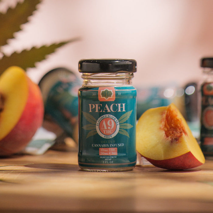 Peach Tea Shot sitting on table with peach fruit and cannabis leaves around it.